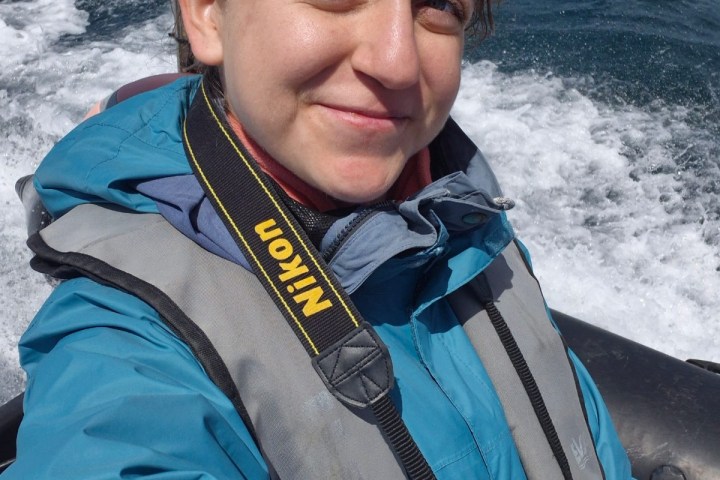 Person in blue jacket and life vest on a boat, holding a camera with ocean in the background.
