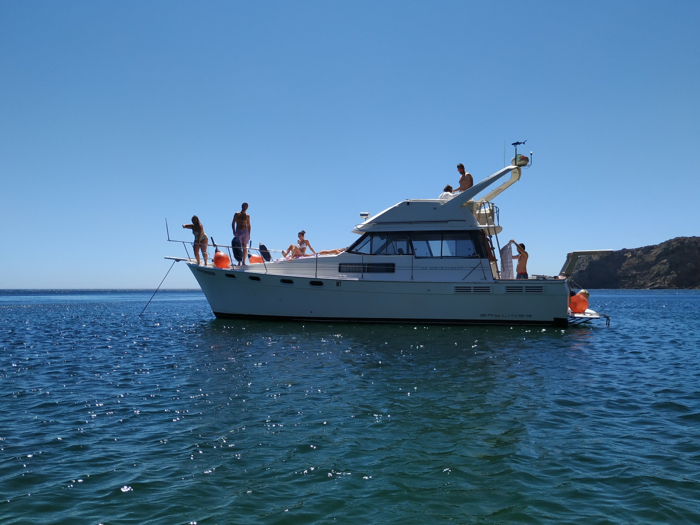People on a yacht in sunny weather on a calm sea with a cliff in the background.