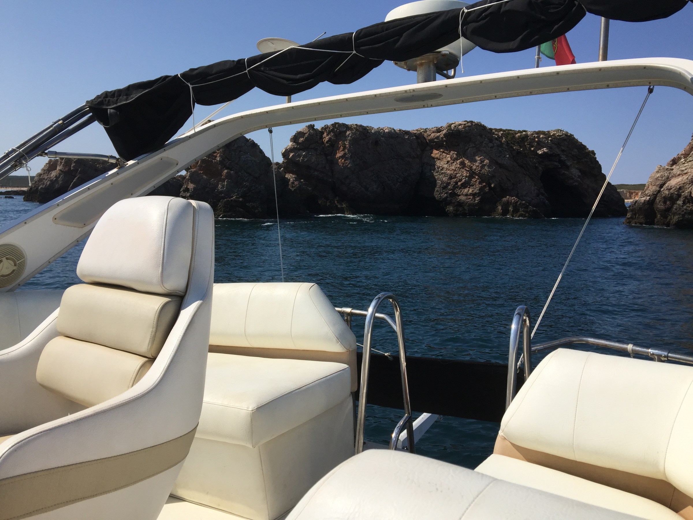 Boat interior with white seats, rocky coastline in background under clear sky.