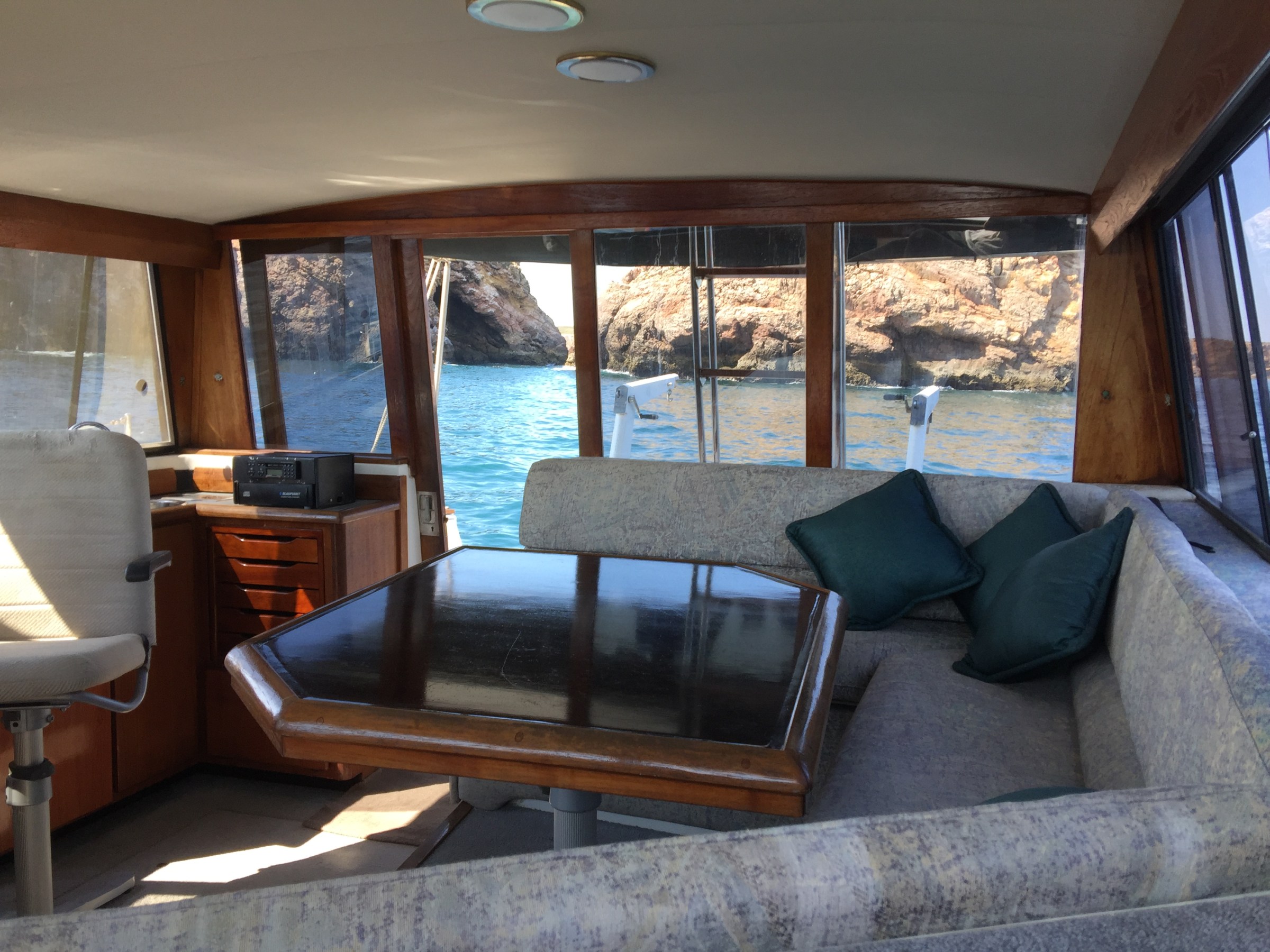 Interior of a boat with a table, chairs, and windows showing rocky coastline and water outside.